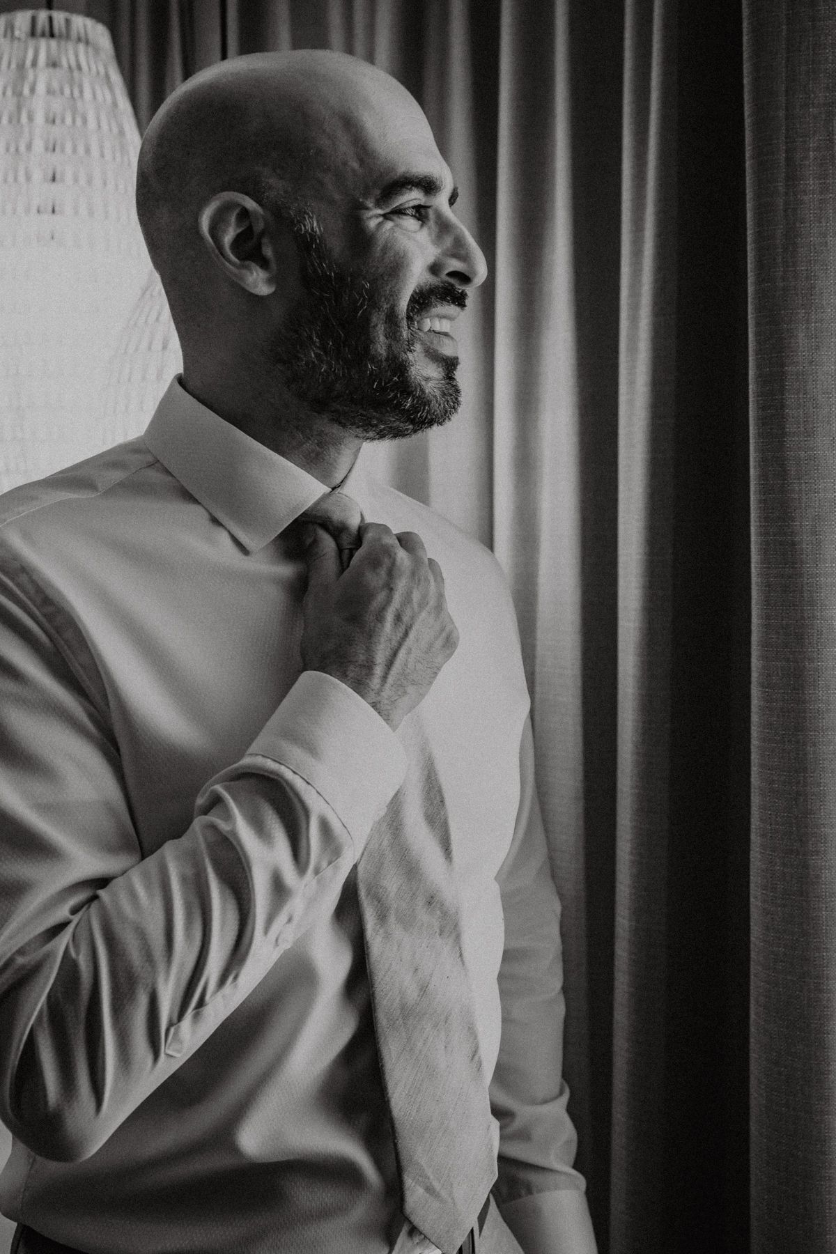 Black and white photo of a bald bearded man adjusting his tie while getting dressed in a dress shirt.