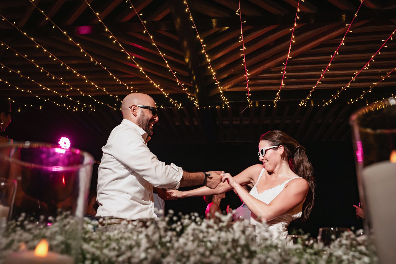 Couple dancing joyfully under string lights at a romantic wedding reception with candles and floral centerpieces.