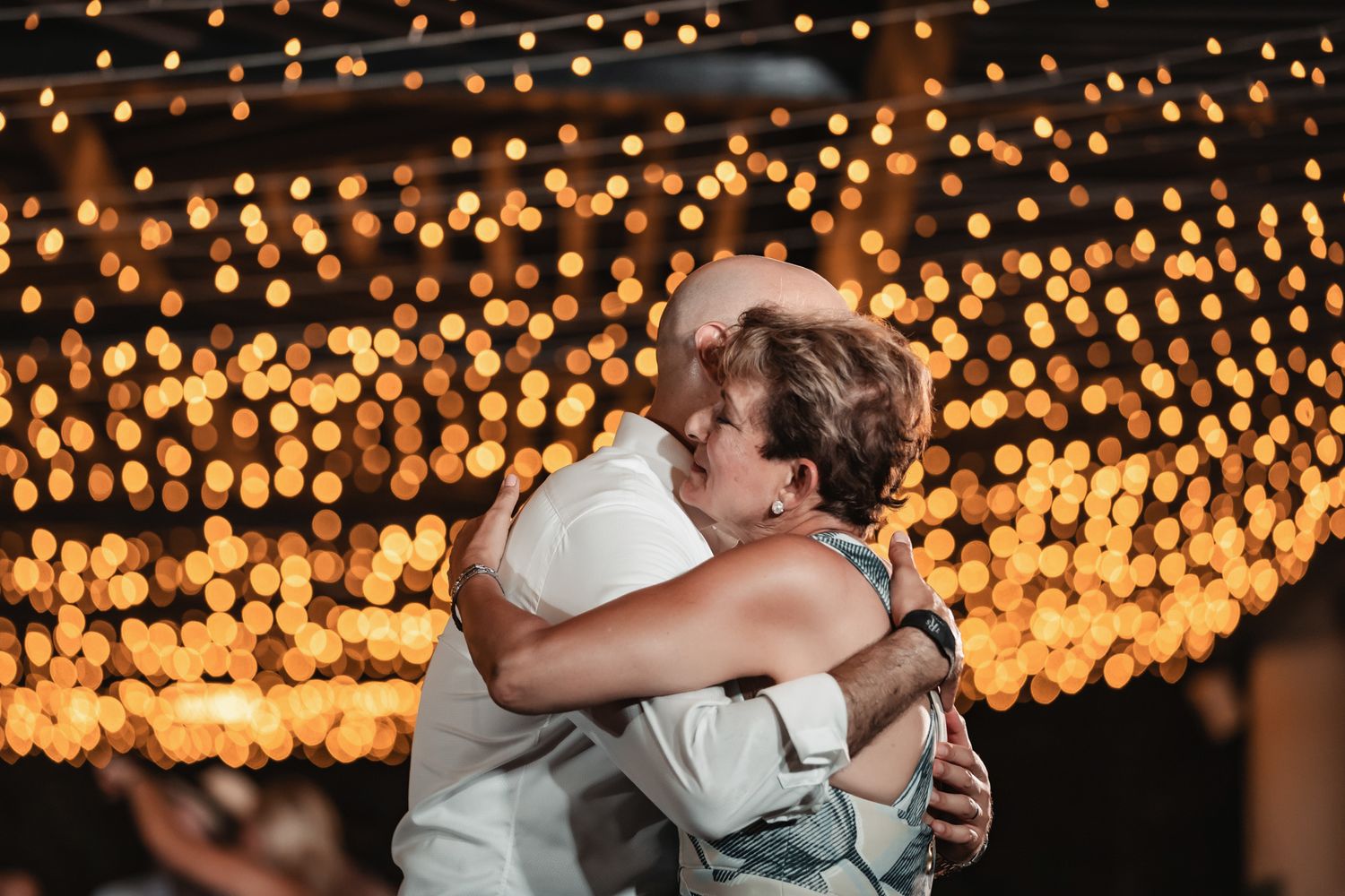 Two people share a warm embrace on a dance floor surrounded by glowing golden bokeh string lights at a wedding reception.