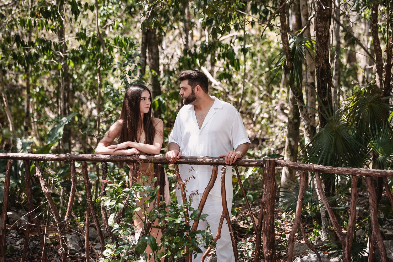 Couple in white attire standing on a rustic wooden bridge surrounded by lush tropical forest greenery.