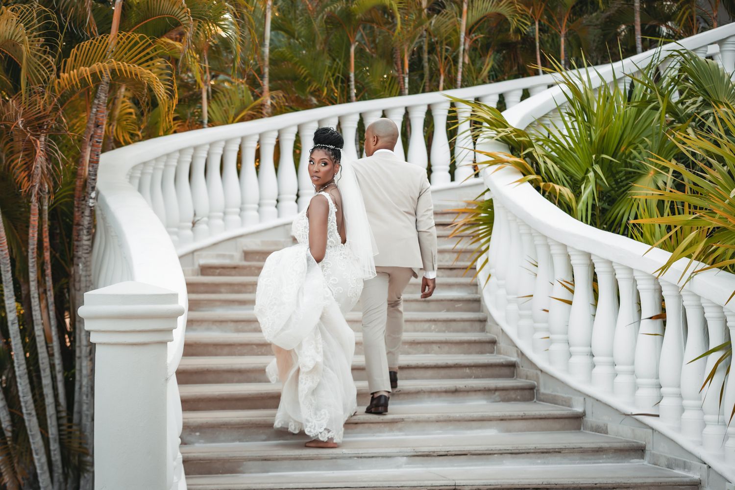 Bride and groom walking up elegant white staircase surrounded by tropical palms at outdoor wedding.