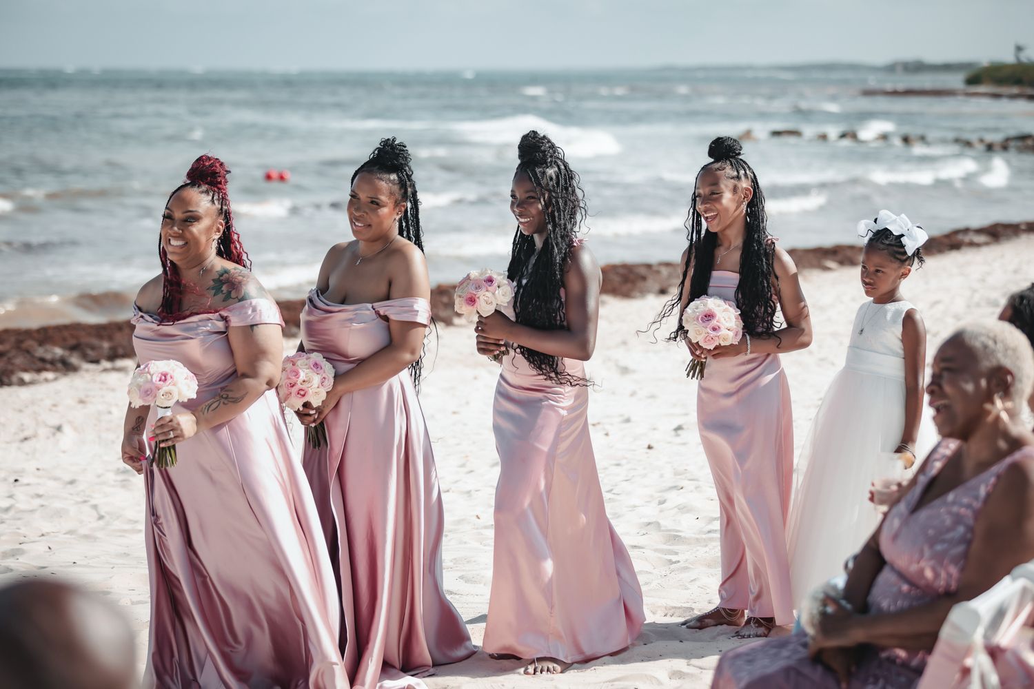Bridesmaids in pink satin dresses holding bouquets stand on a beach during an outdoor wedding ceremony.