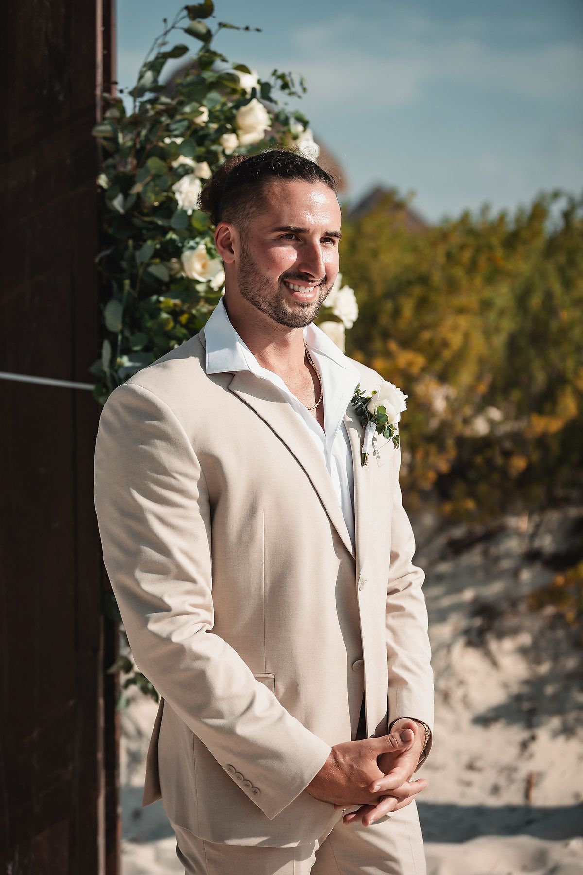 Smiling groom in beige suit stands by floral arch on sandy beach at outdoor wedding ceremony.