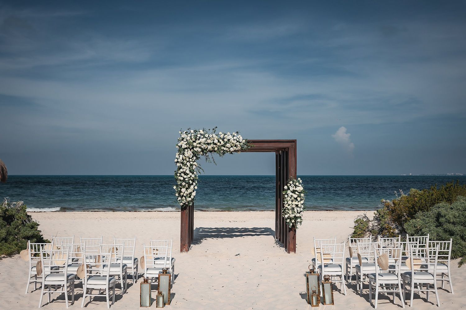 Beach wedding ceremony setup with white floral arch, white chairs, and ocean backdrop under blue sky.