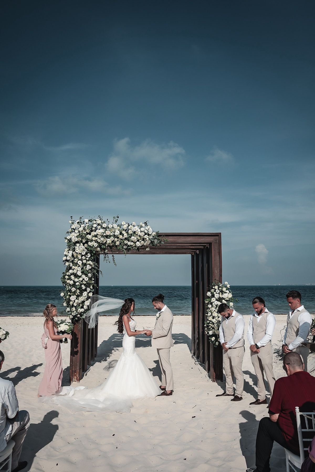 Couple exchanging vows at a beach wedding ceremony under a floral arch with white flowers and ocean backdrop.