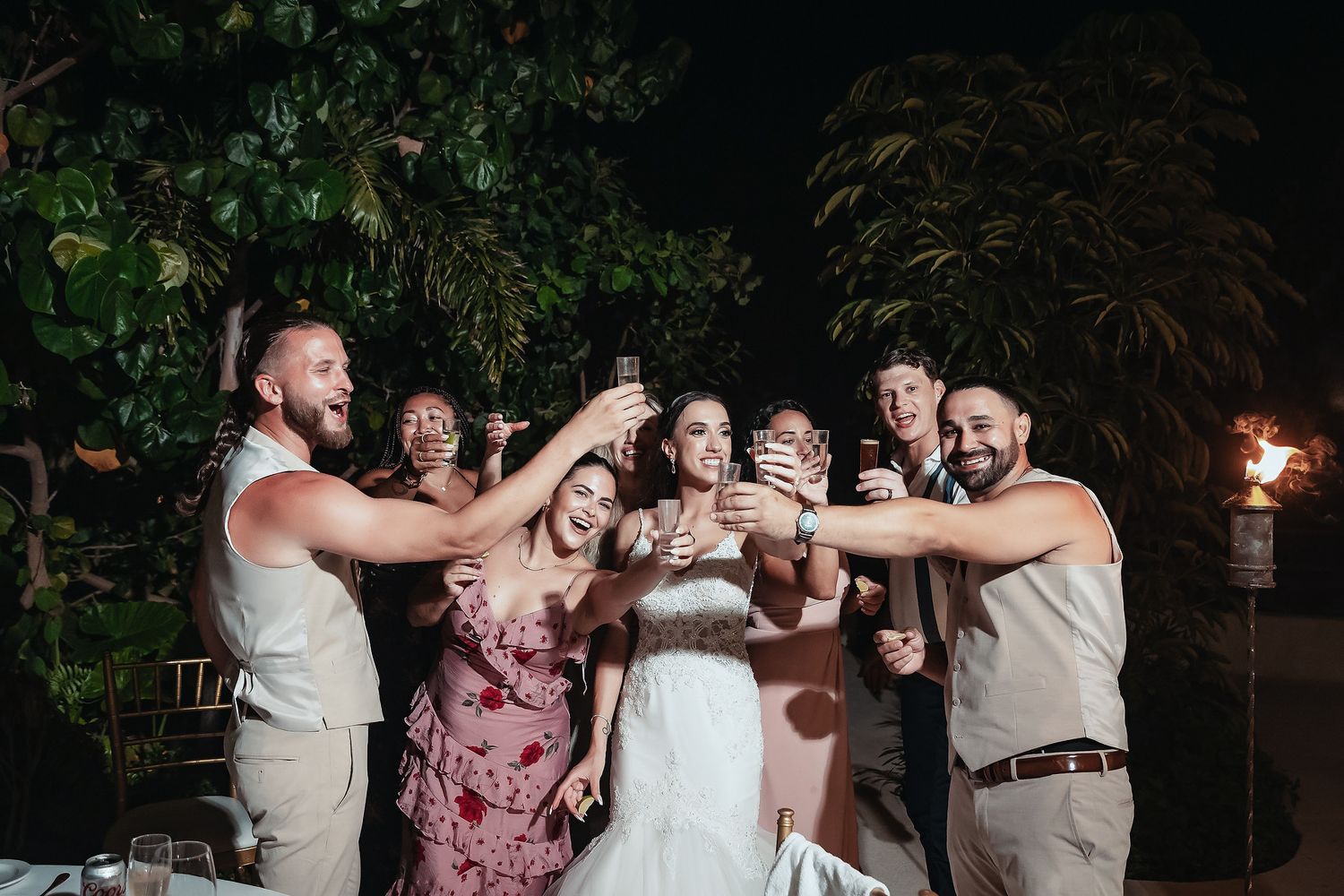 Wedding party toasting with drinks outdoors at night, bride in white gown surrounded by smiling guests.