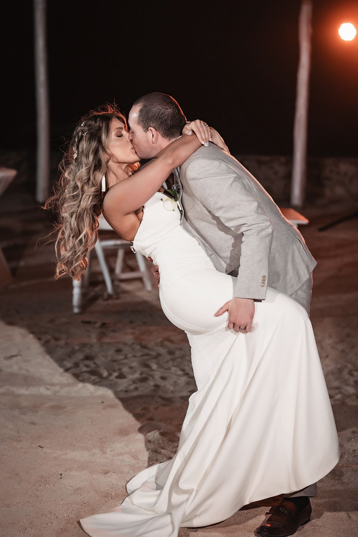 Groom dips bride for a romantic kiss on the beach at night during their outdoor wedding reception.