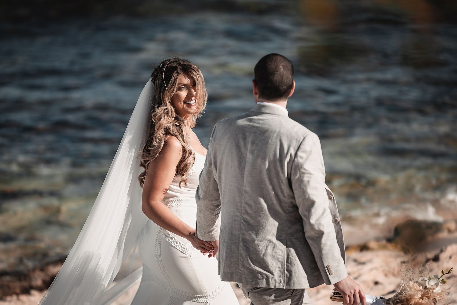 Bride in white gown and veil holds hands with groom in gray suit by a rocky shoreline.