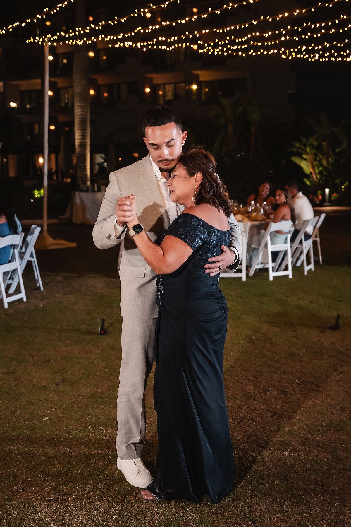 Groom in tan suit shares mother-son dance at outdoor wedding reception with string lights at night.
