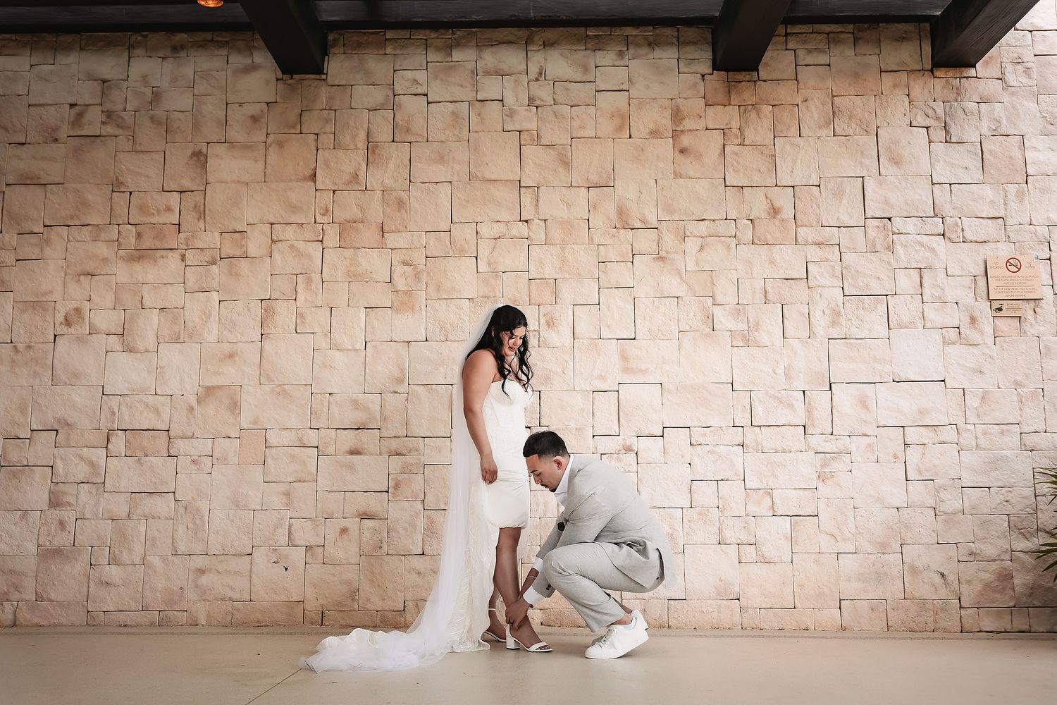 Groom kneeling to adjust bride's shoes against a textured stone wall in an elegant wedding venue.
