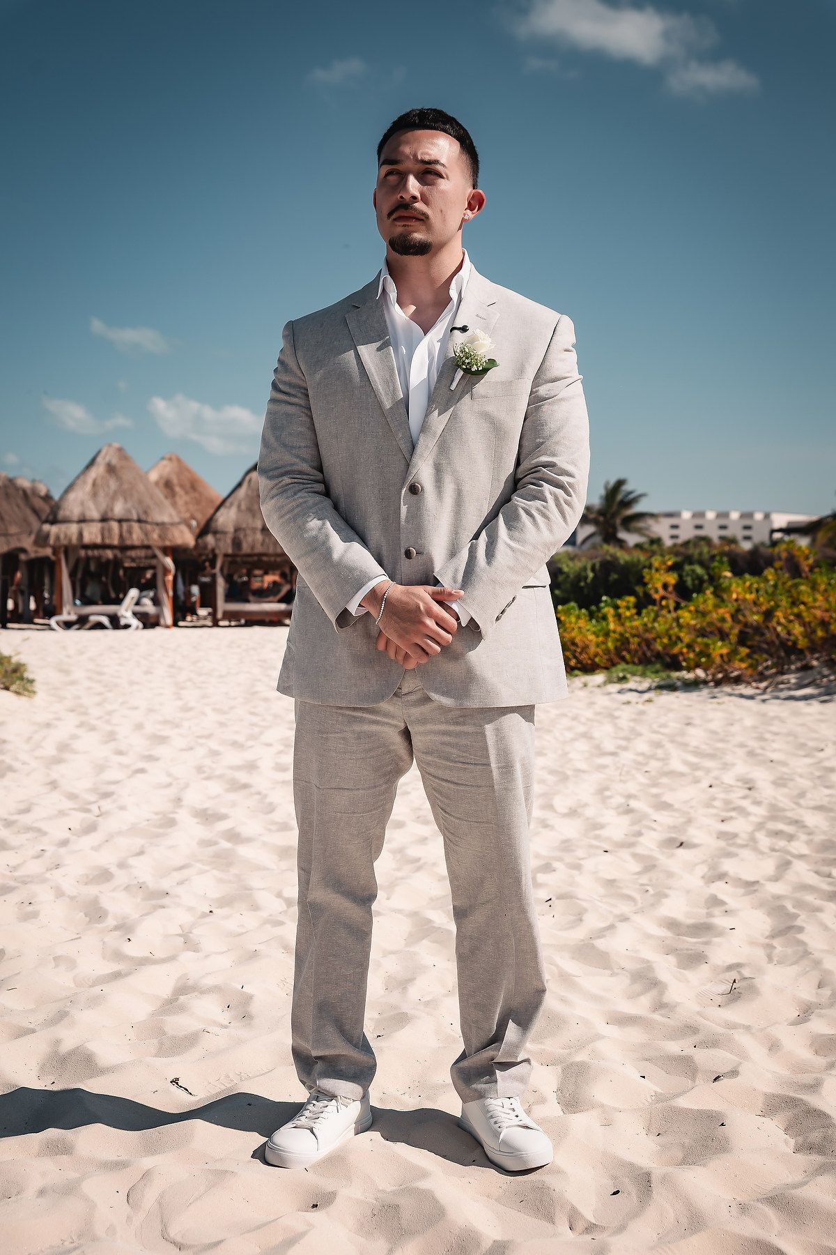 Groom in a light gray linen suit with boutonniere and white sneakers standing on a sandy beach.