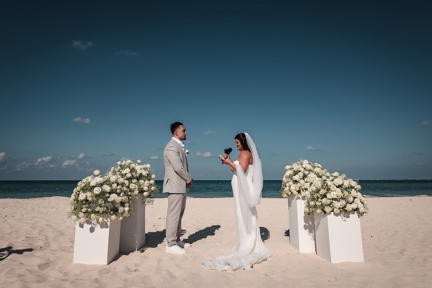 Couple exchanges vows at a romantic beach wedding ceremony with white floral arrangements under a clear blue sky.
