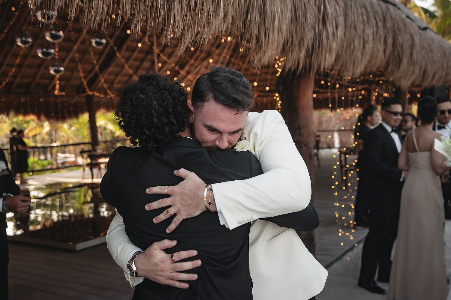 Groom in white tuxedo shares heartfelt hug with guest at tropical outdoor wedding reception venue.