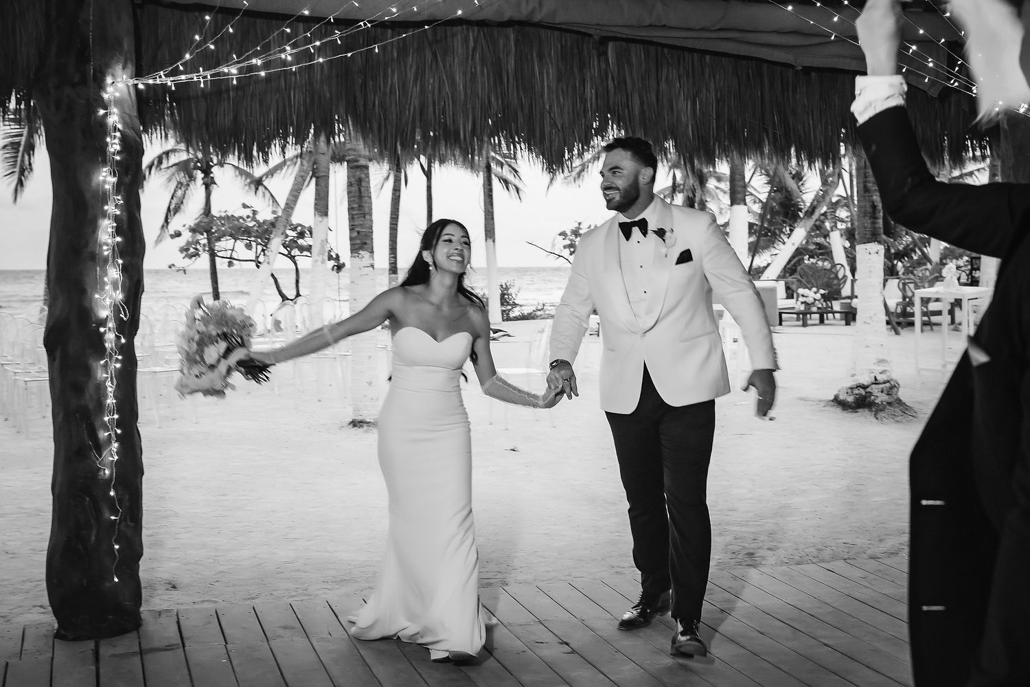 Newlyweds walk hand in hand under a thatched roof on a sandy beach, celebrating their tropical wedding.