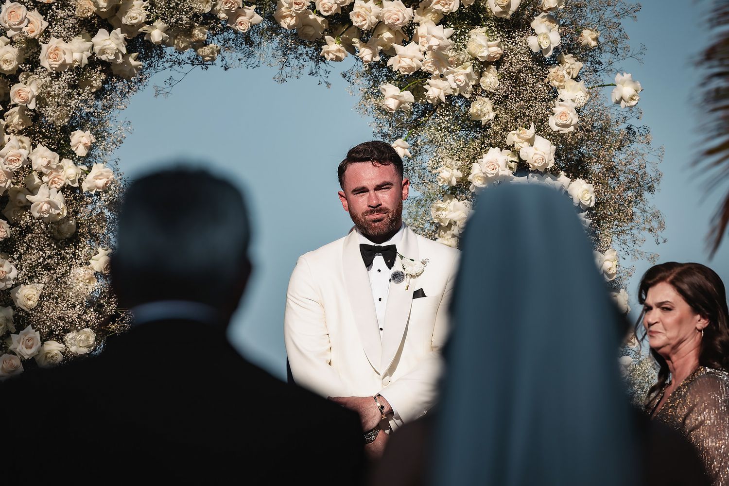 Groom in white tuxedo stands at floral arch altar during outdoor wedding ceremony.