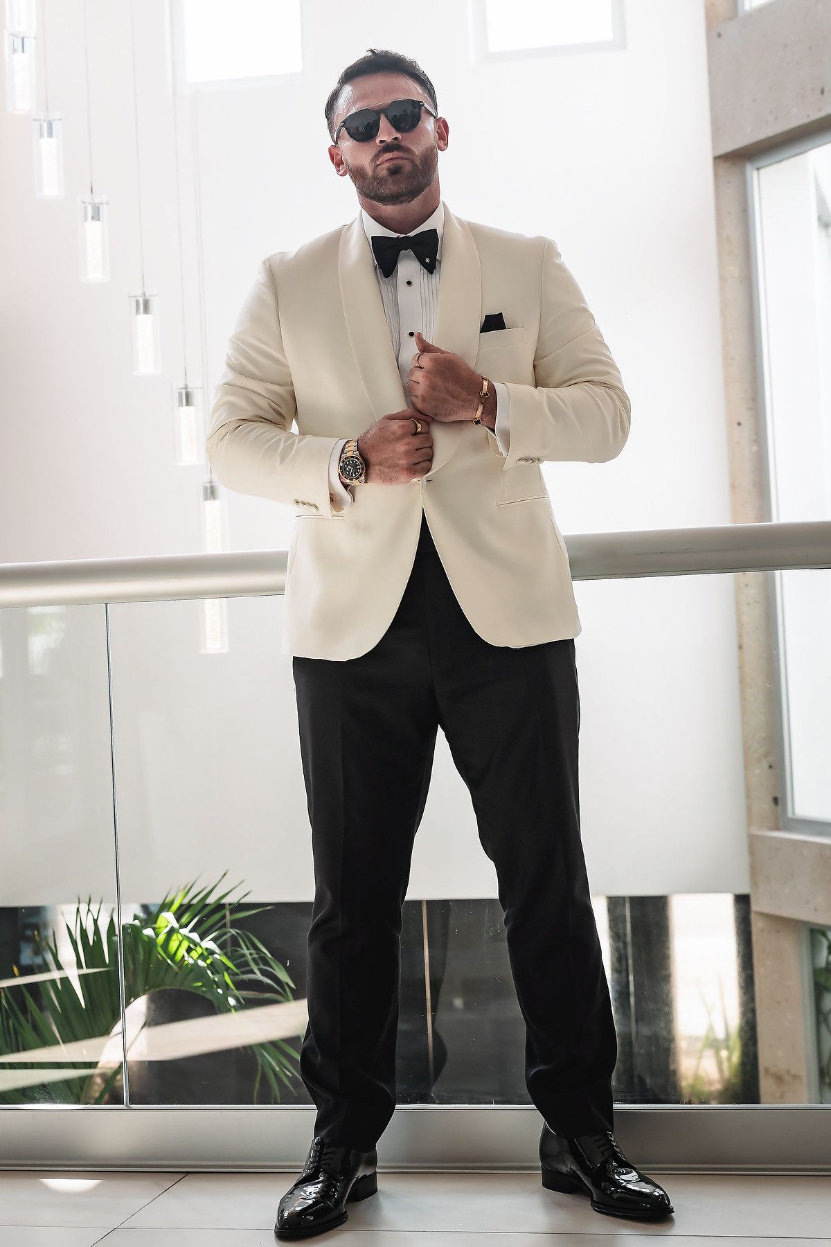 Man in cream tuxedo jacket, black bow tie, trousers, and sunglasses posing confidently in a modern interior setting.