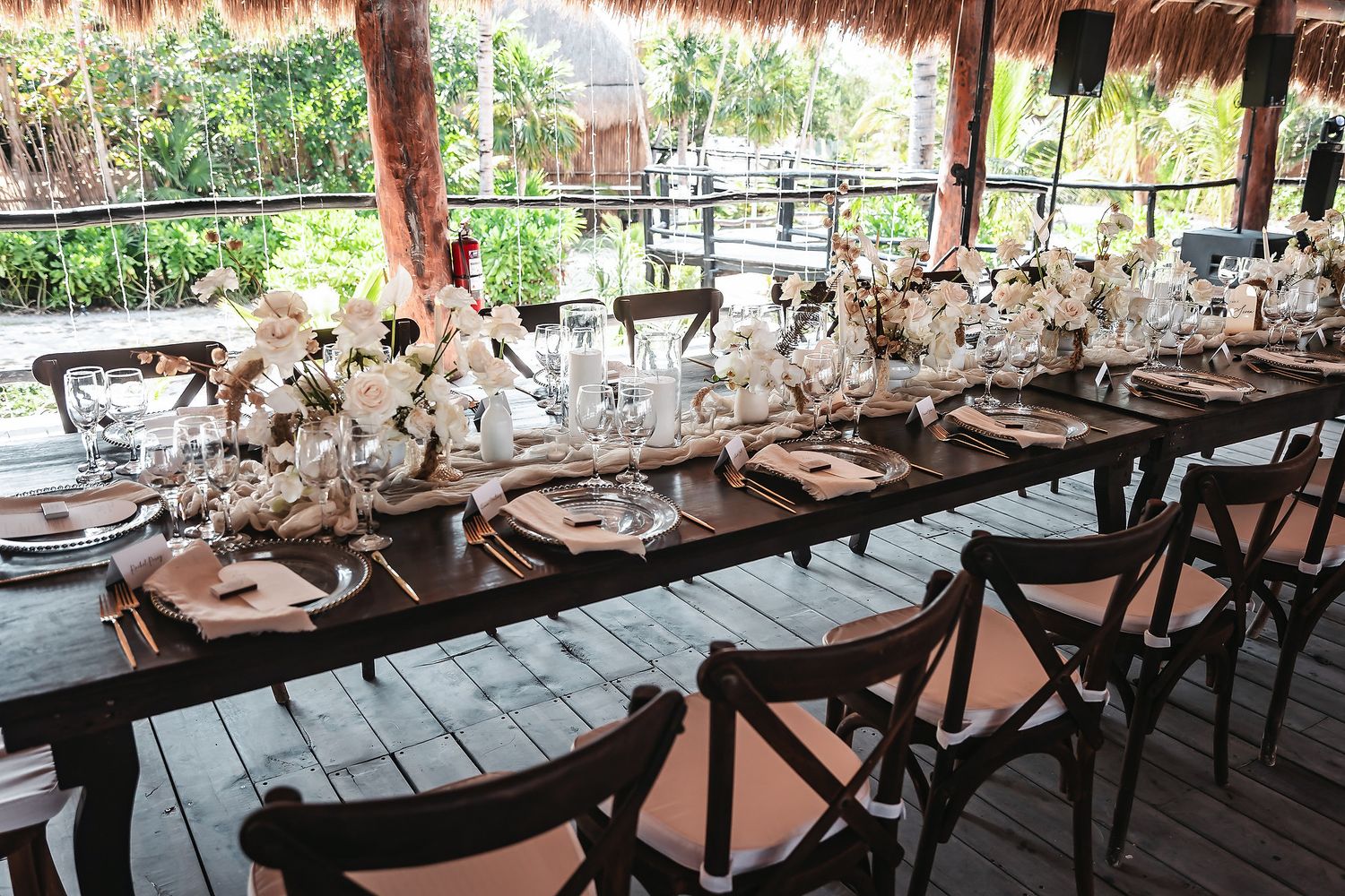 Elegant outdoor wedding reception table with white floral centerpieces, wooden chairs, and tropical greenery backdrop.