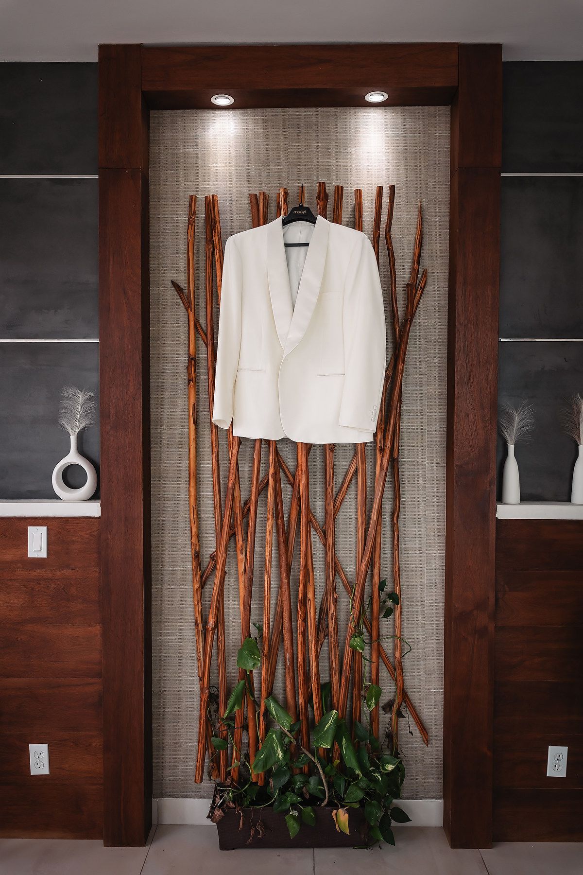 White tuxedo jacket displayed on decorative wooden branches in an illuminated niche with green plants below.