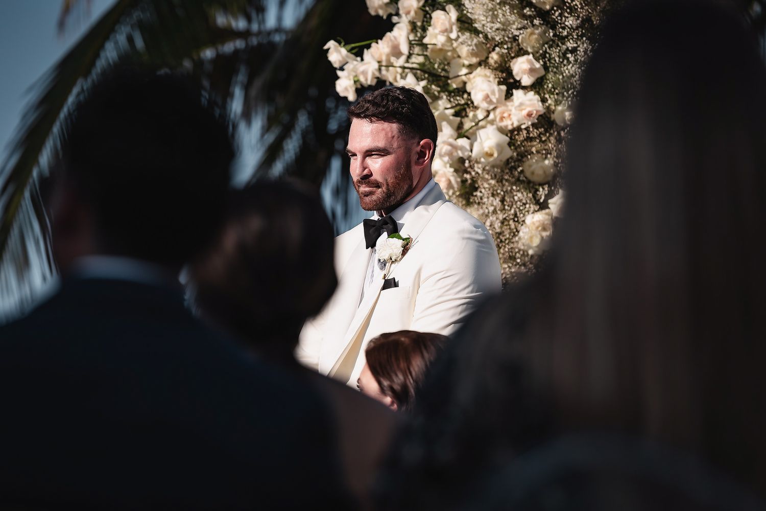 Groom in white tuxedo standing at outdoor wedding altar surrounded by white floral arrangements and guests.