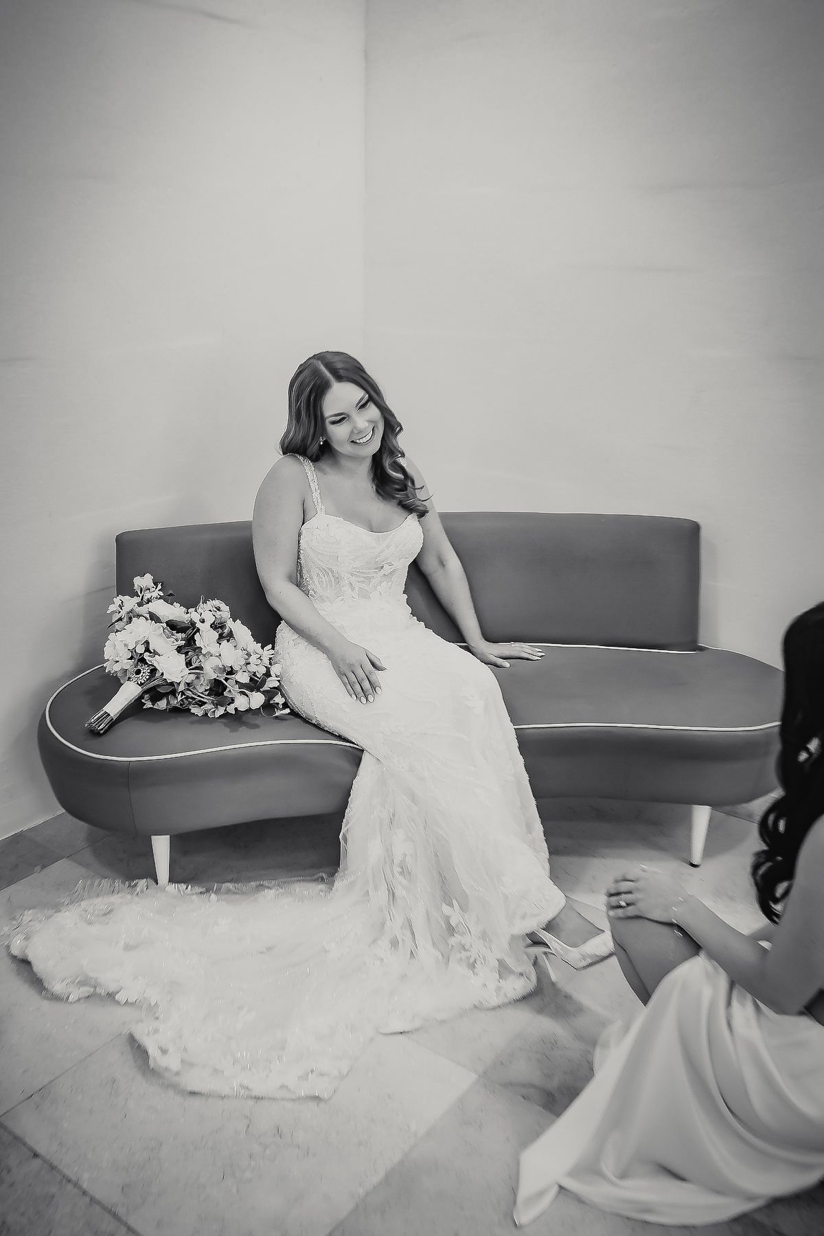 Bride in lace gown sits on sofa smiling with bouquet beside her in elegant black and white photo.