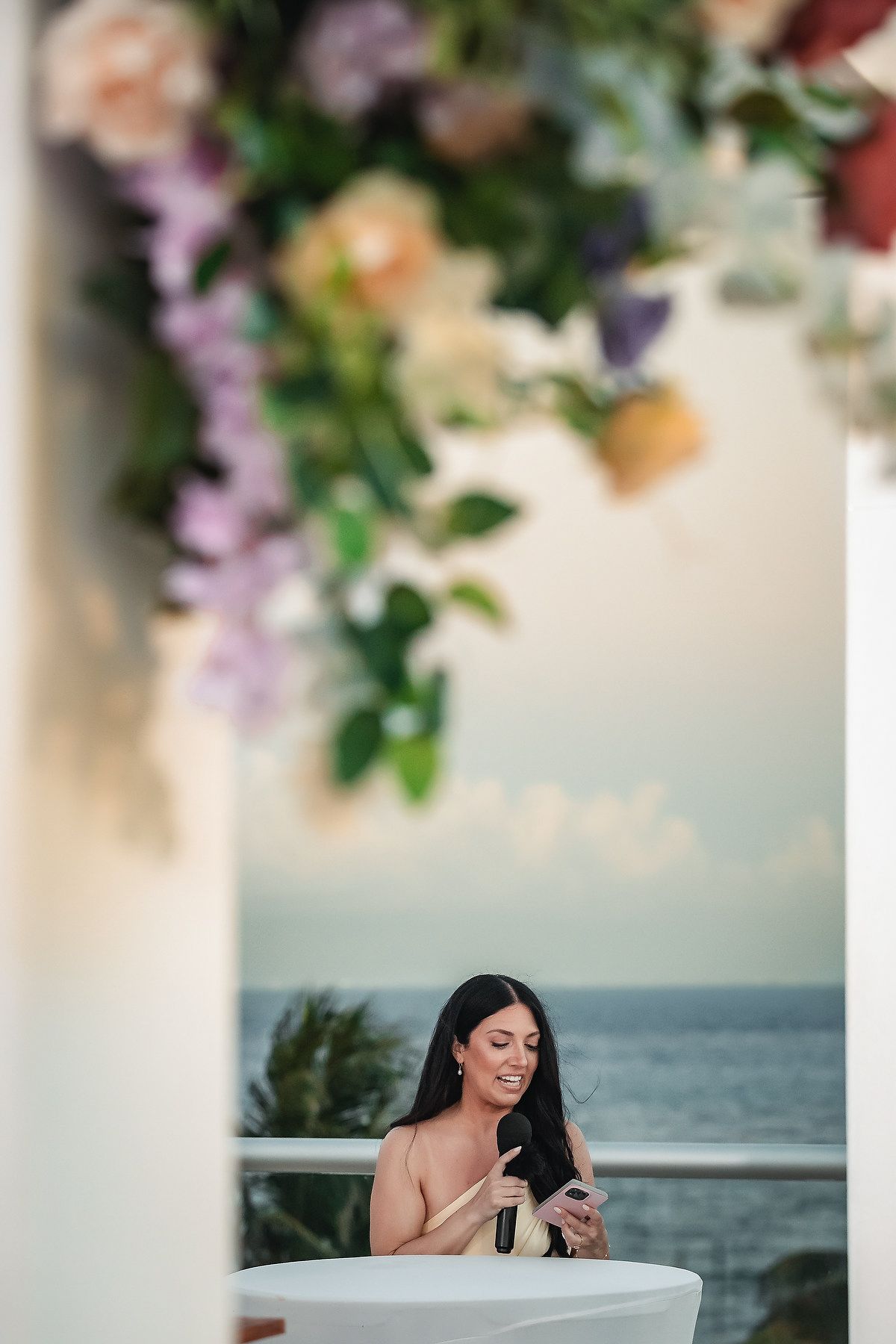 Smiling brunette woman in elegant attire holds a drink on an oceanview balcony adorned with colorful floral arrangements.