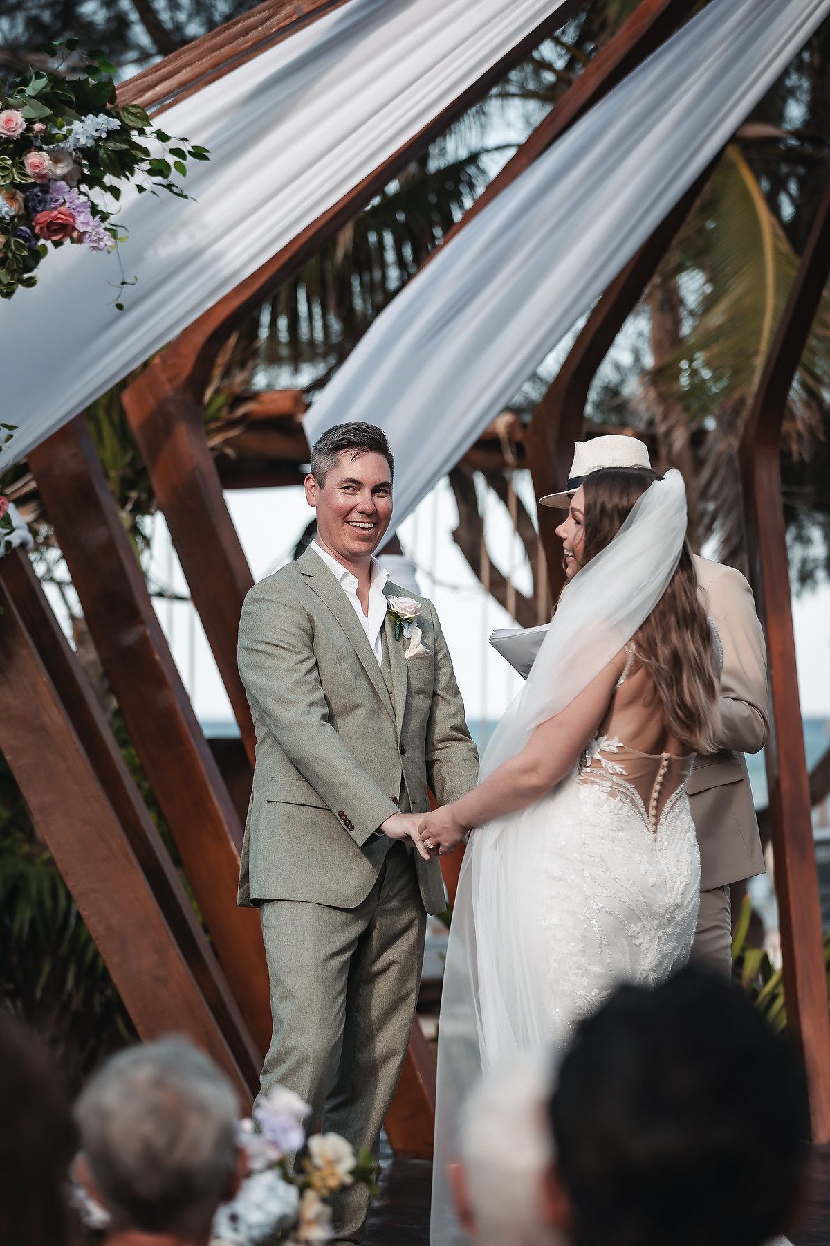Couple holding hands at outdoor wedding ceremony under geometric wooden arch with white draping and floral decor.