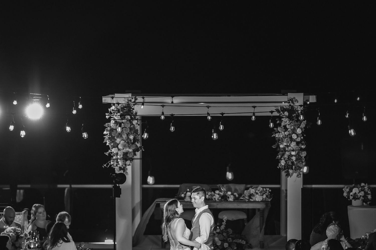 Couple embracing under floral arch with string lights at outdoor night wedding ceremony in black and white photo.