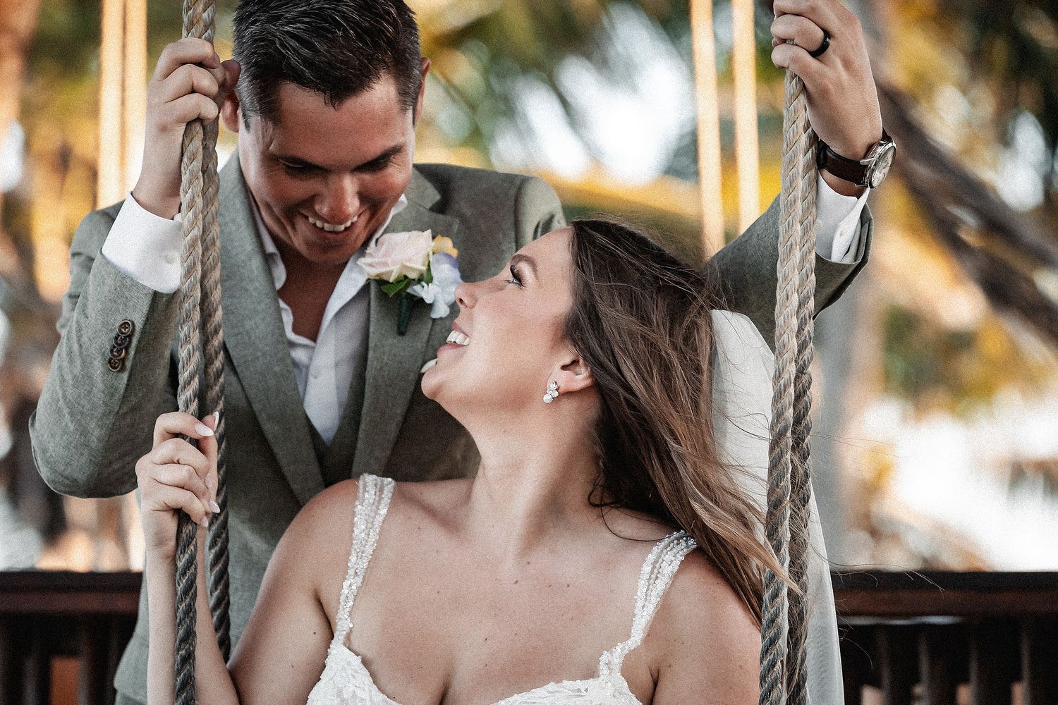 A joyful bride and groom share a romantic moment on a swing, surrounded by tropical palms on their wedding day.
