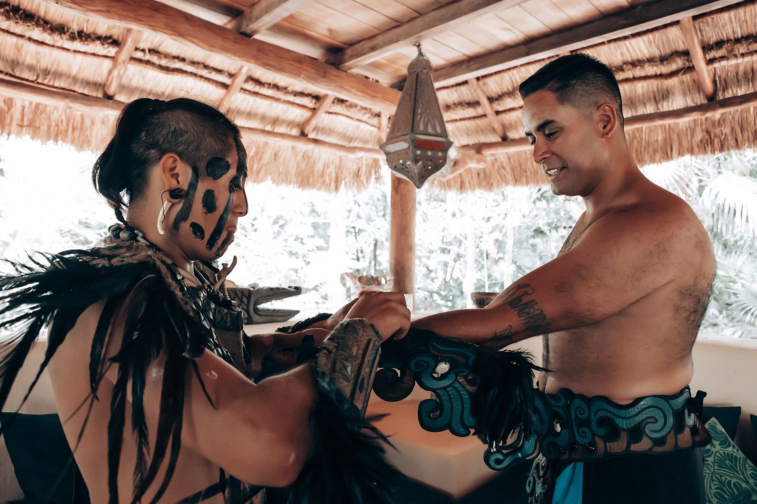 Two performers in traditional Mayan costumes with face paint and feathered regalia engage in a ceremonial ritual indoors.