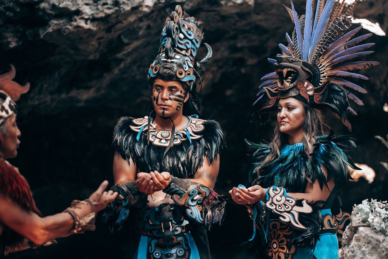 Two performers in detailed blue Mayan ceremonial feathered costumes holding masks in a cave.