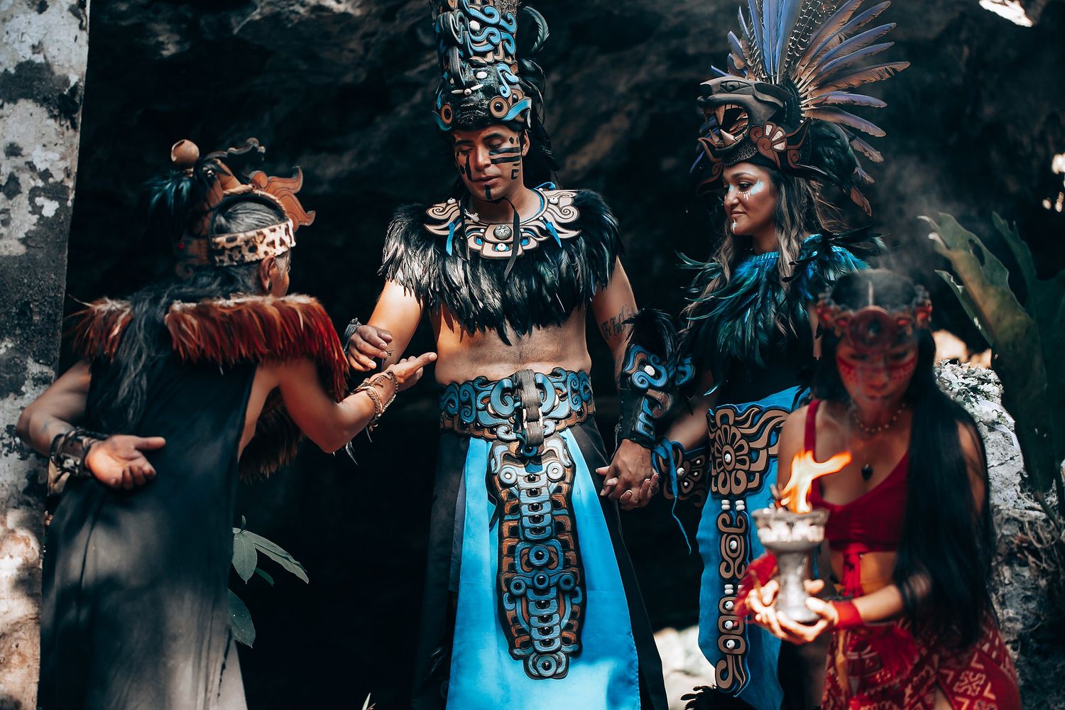 Group of four performers in elaborate blue and black Mayan ceremonial costumes holding candles.