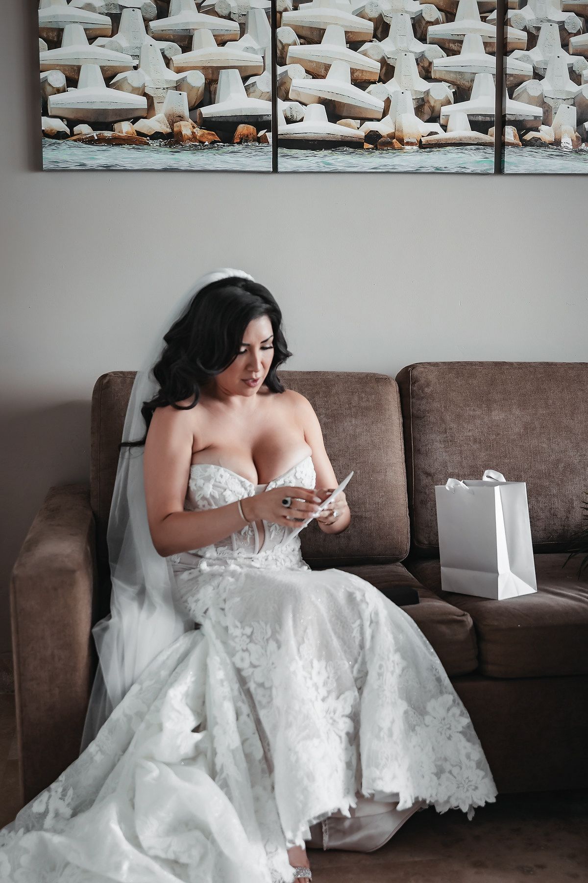 Bride in white lace wedding gown and veil sits on brown sofa reading a note, with gift bag beside her.