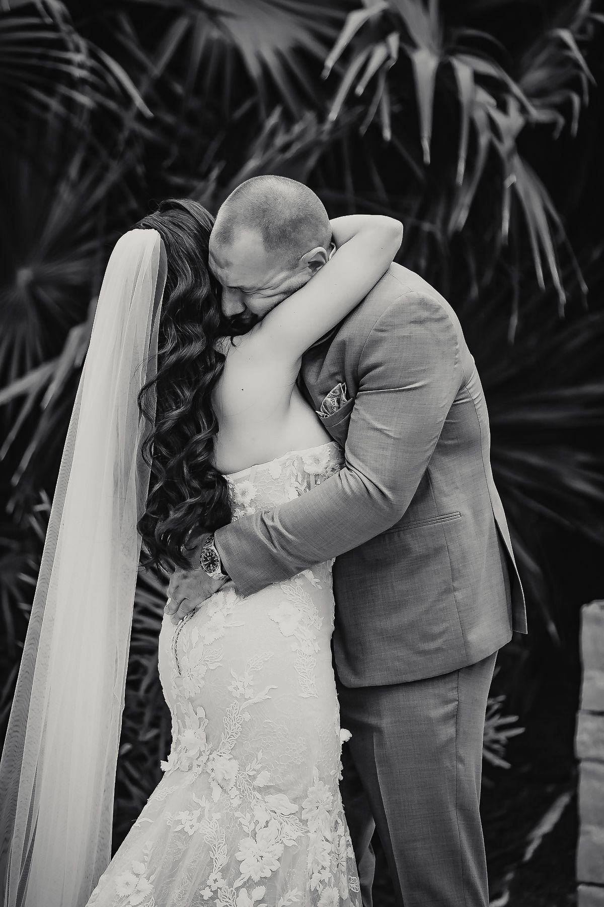 Black and white photo of bride and groom sharing first kiss, bride in lace mermaid gown with veil, tropical palm backdrop.