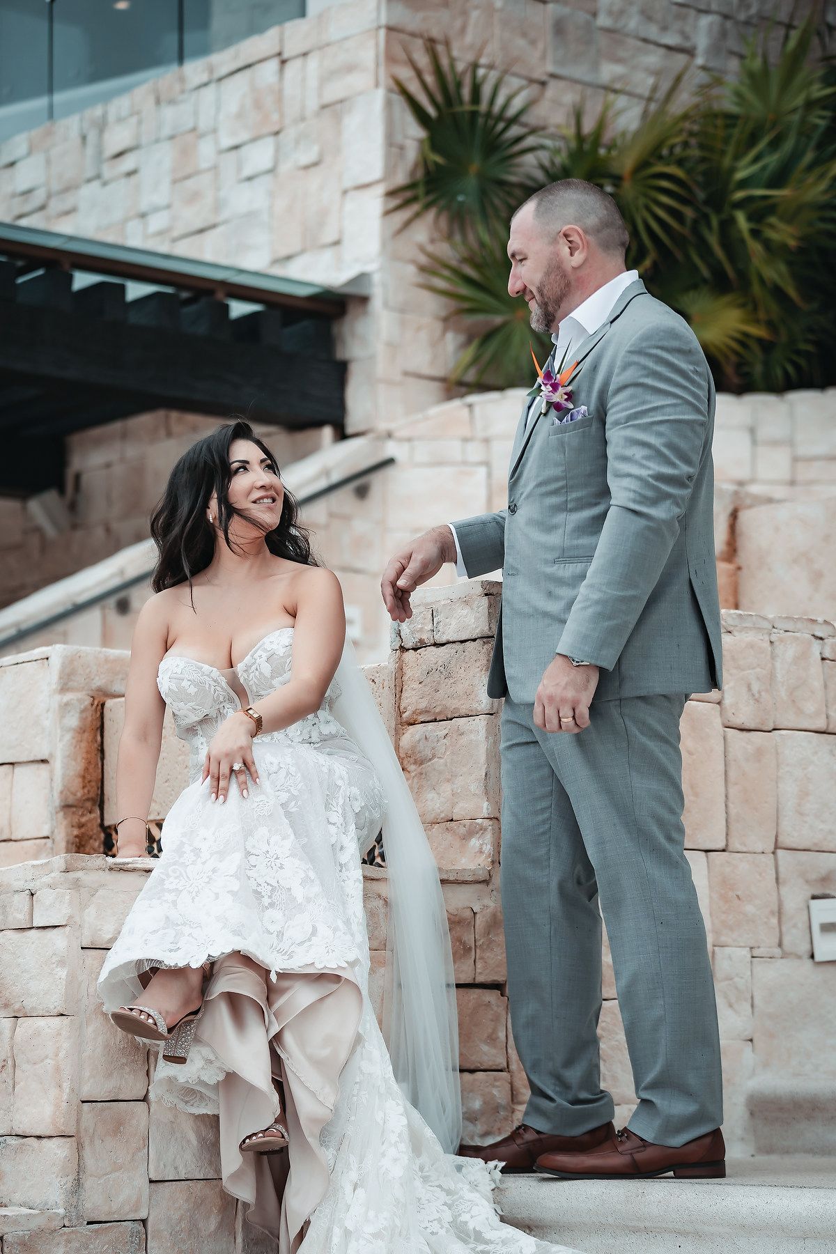 Bride in lace gown sits on stone steps smiling at groom in gray suit at outdoor wedding venue.
