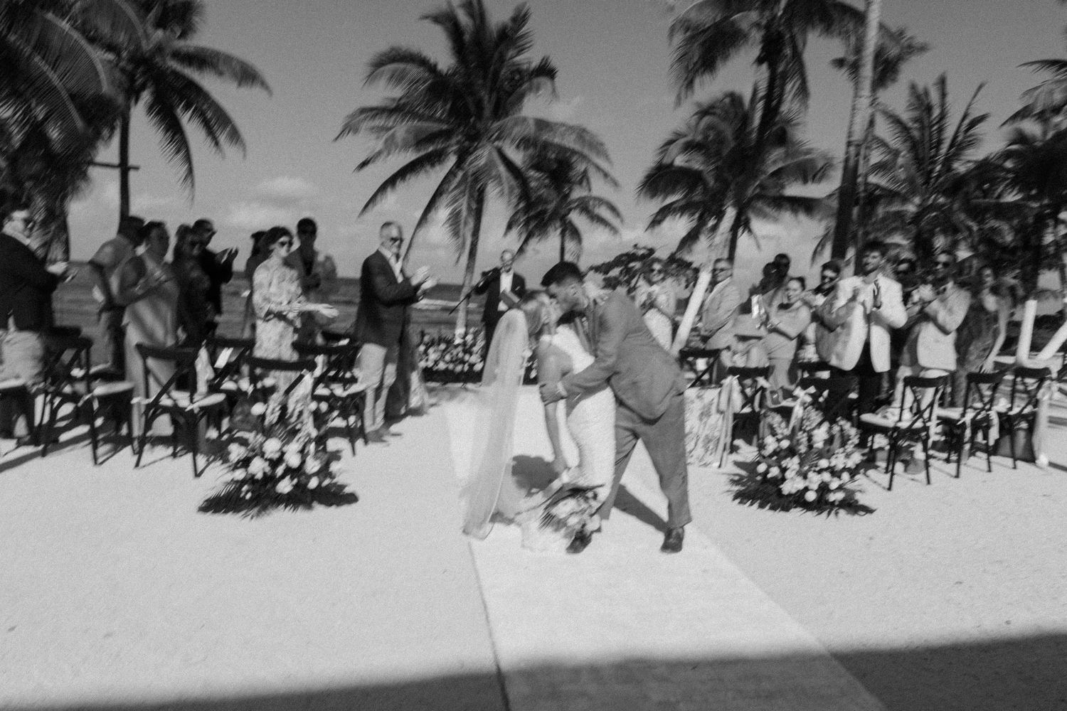 Black and white beach wedding ceremony with couple kissing at altar, palm trees and seated guests in tropical setting.