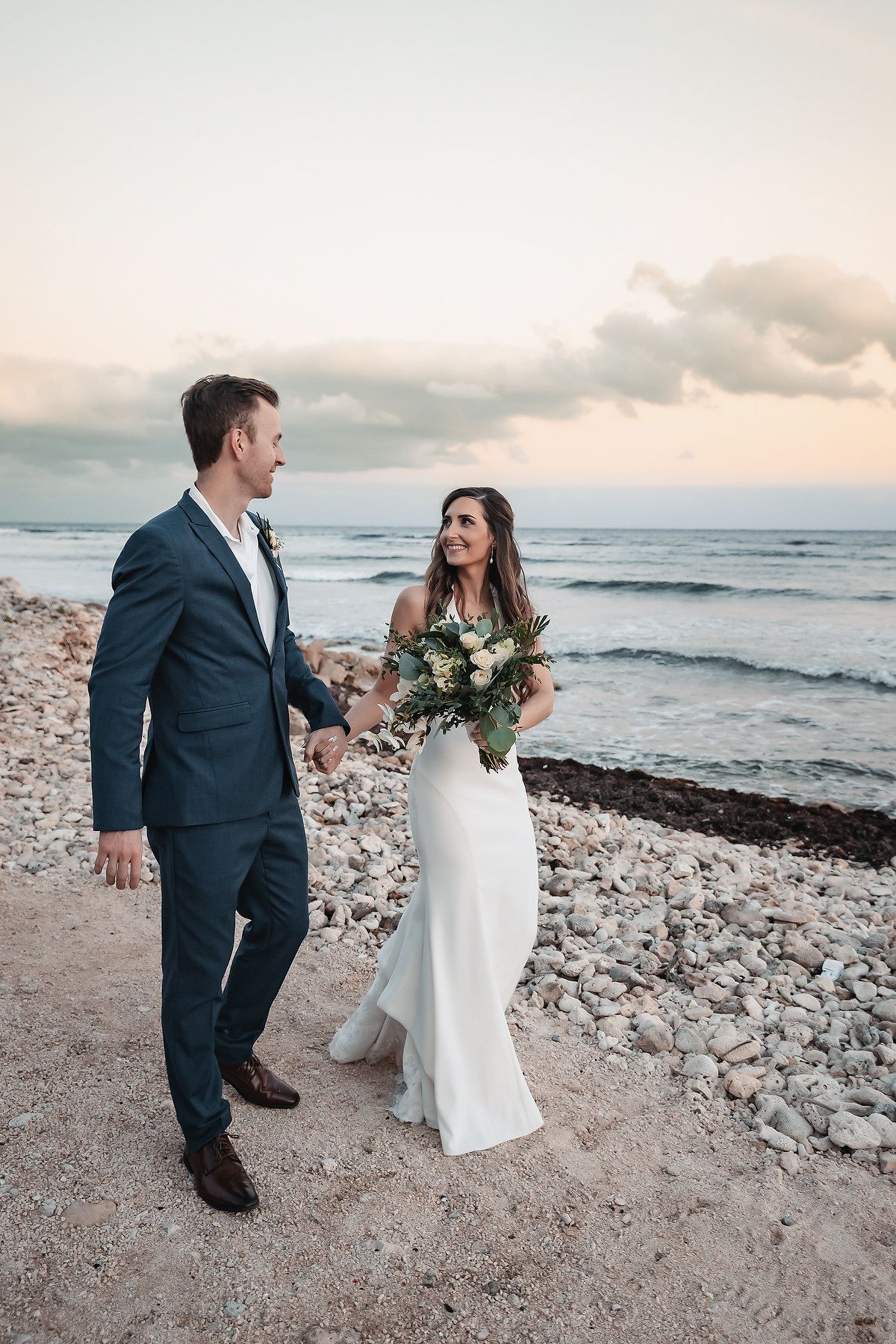 Bride and groom walk hand-in-hand on a rocky beach at sunset, she holds a bouquet in a white gown, he wears a navy suit.