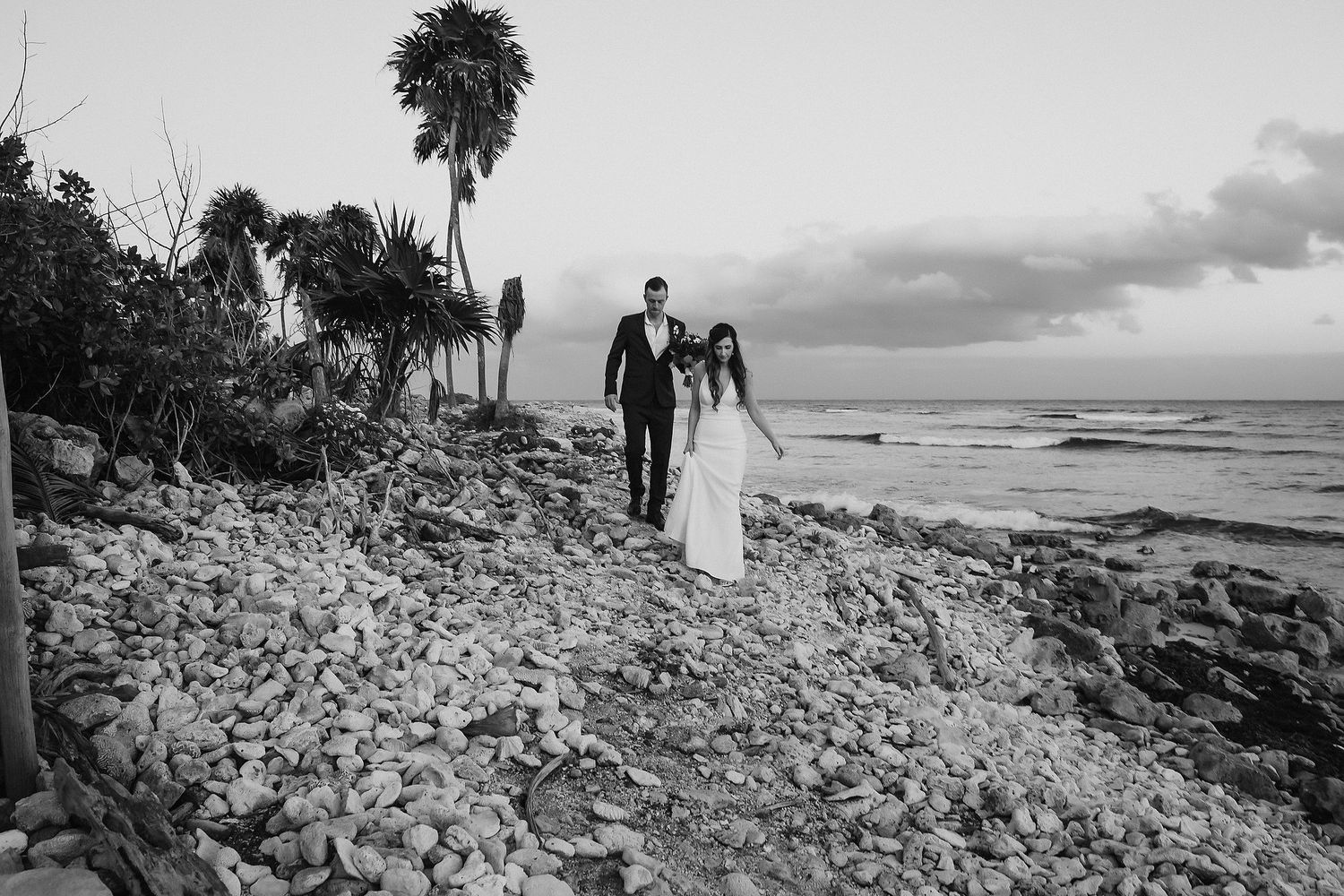 Black and white photo of bride and groom walking hand in hand on a rocky shoreline at dusk.