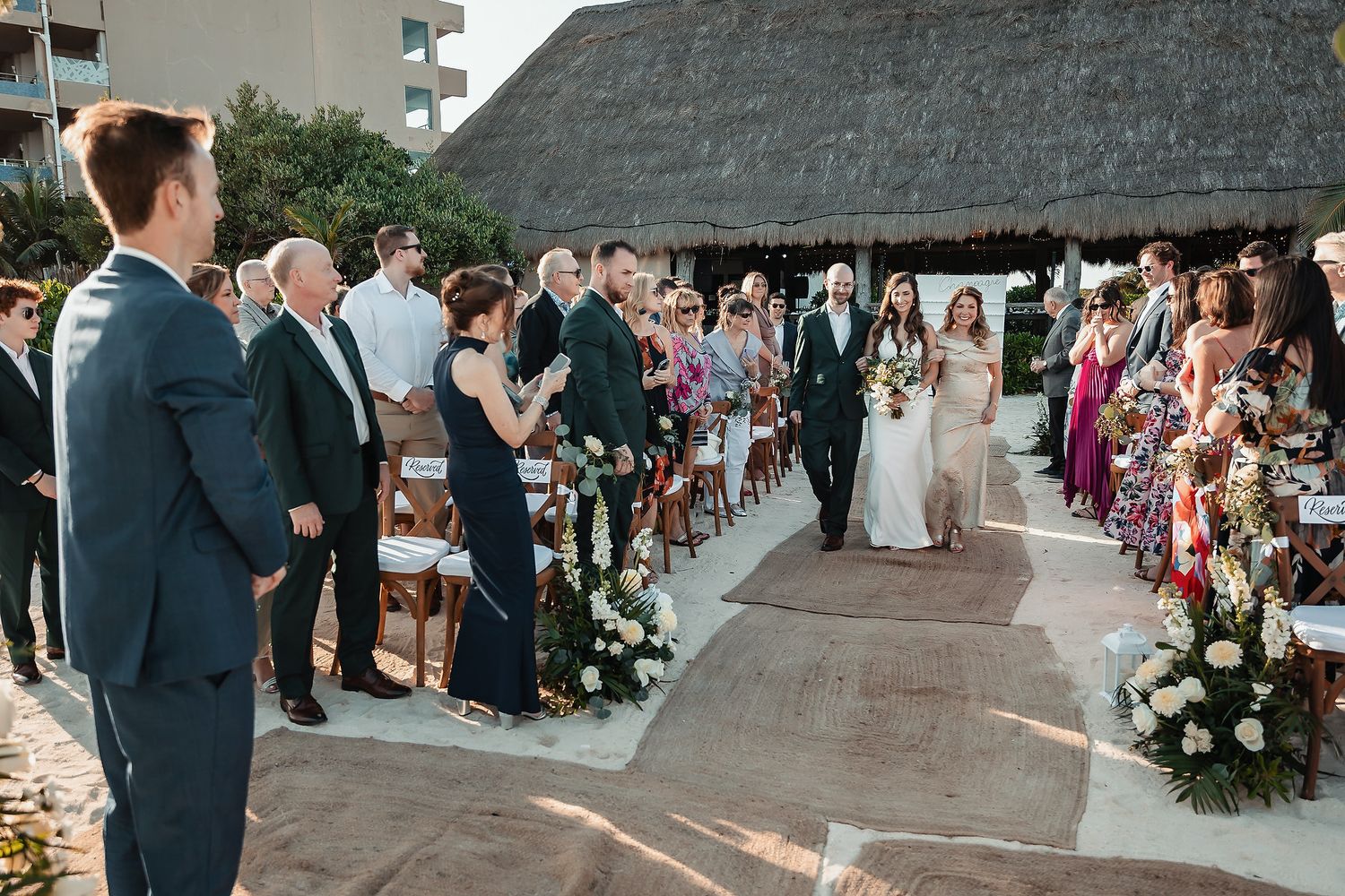 Bride walks down the aisle with escort at outdoor beach wedding ceremony with floral arrangements and guests standing.