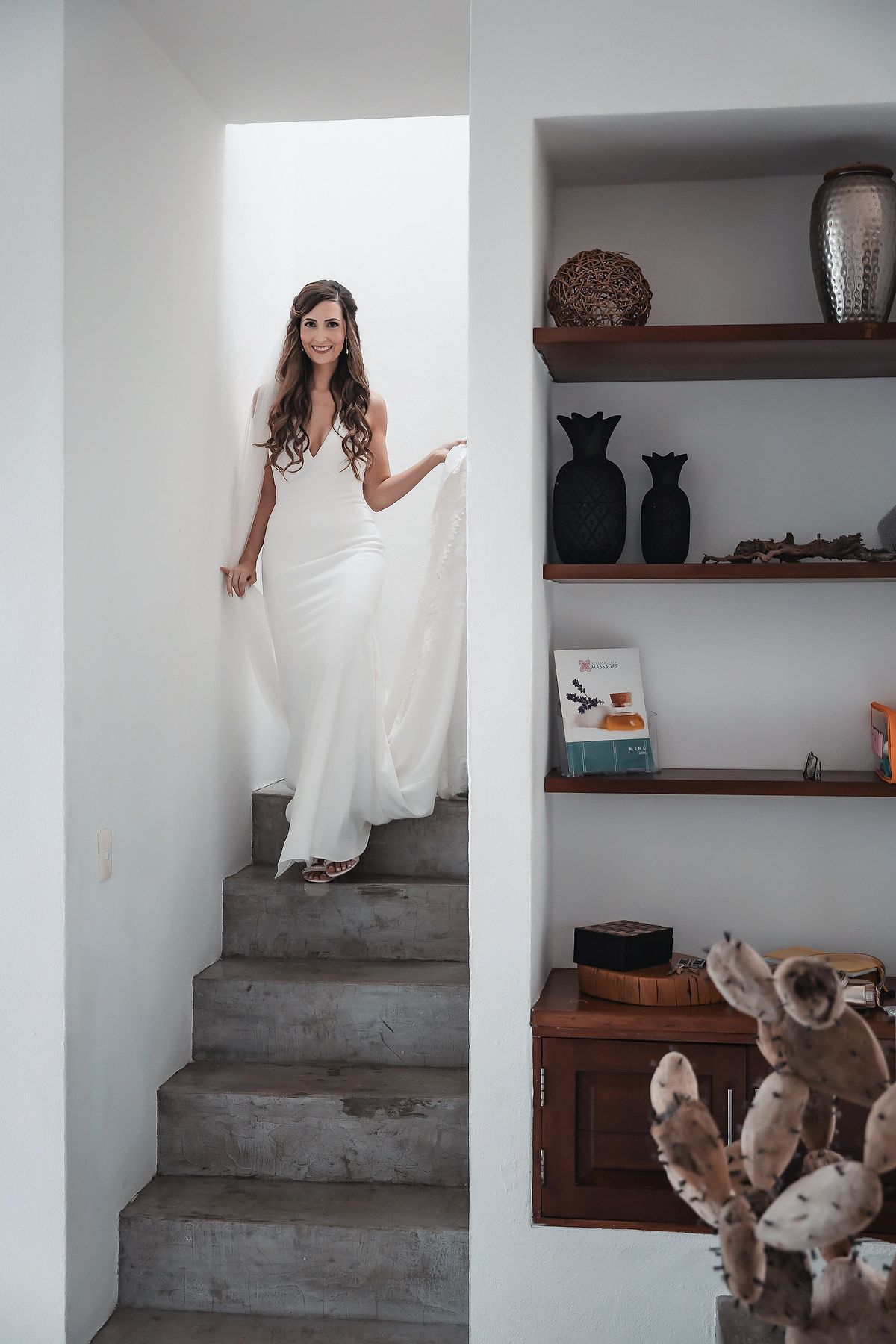 Bride in a flowing white gown descending stone stairs in a modern white interior with wooden shelves and decor.
