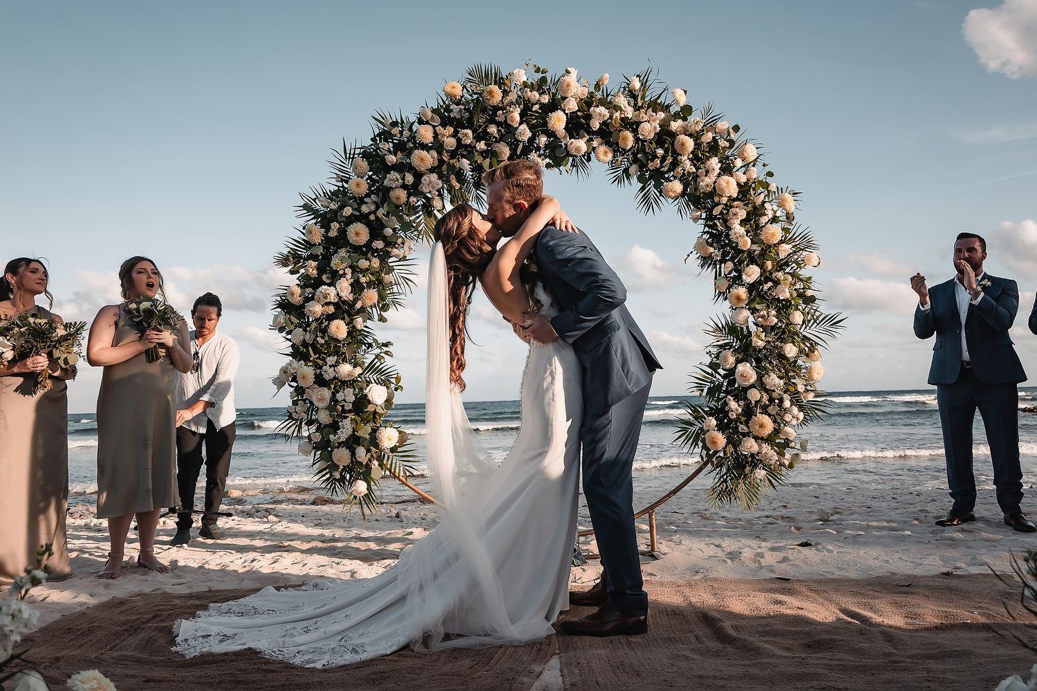 Couple sharing a first kiss under a floral arch at a beach wedding ceremony with guests applauding.