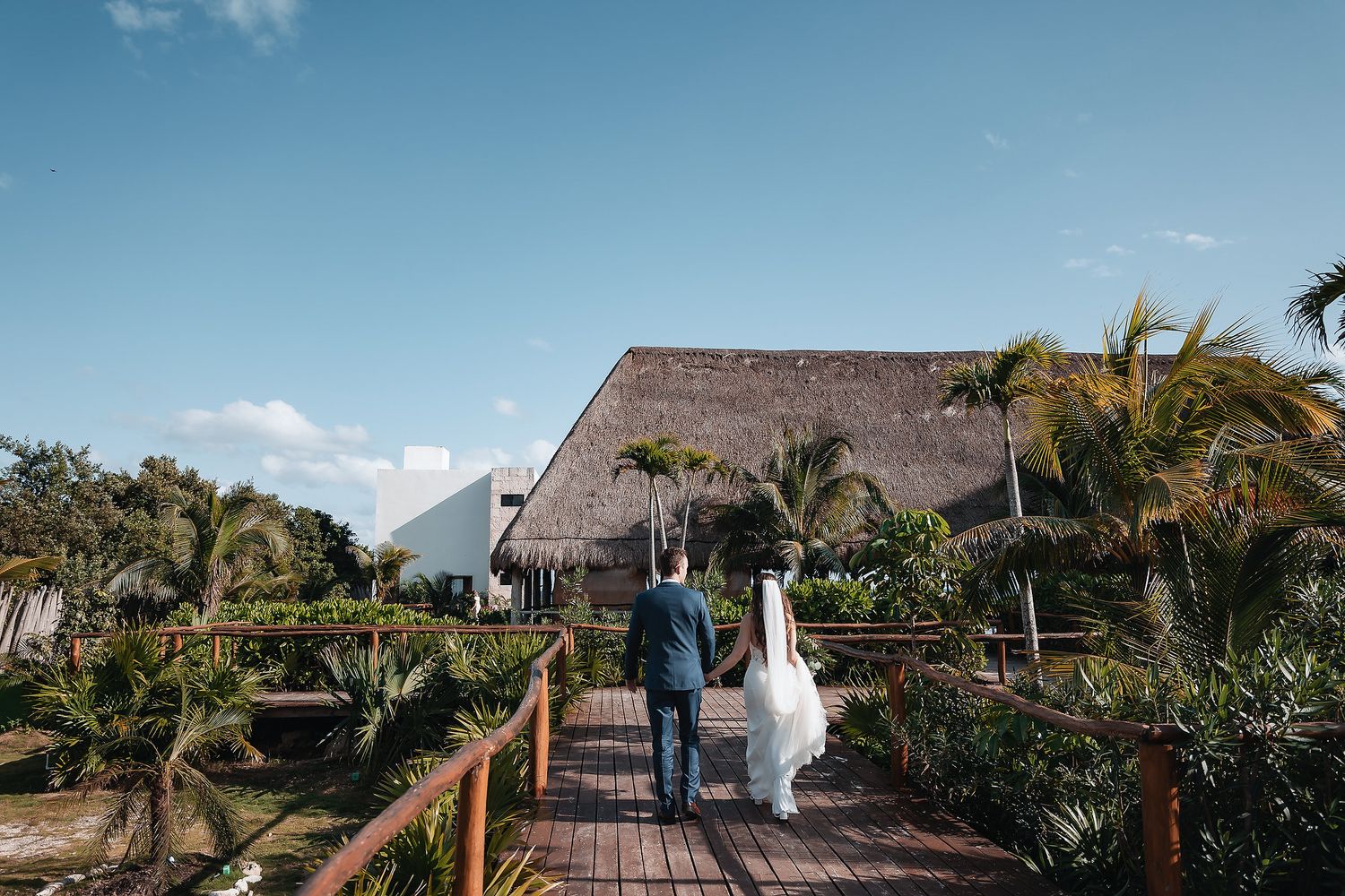 Bride and groom holding hands on a wooden walkway surrounded by tropical palms at a destination wedding venue.