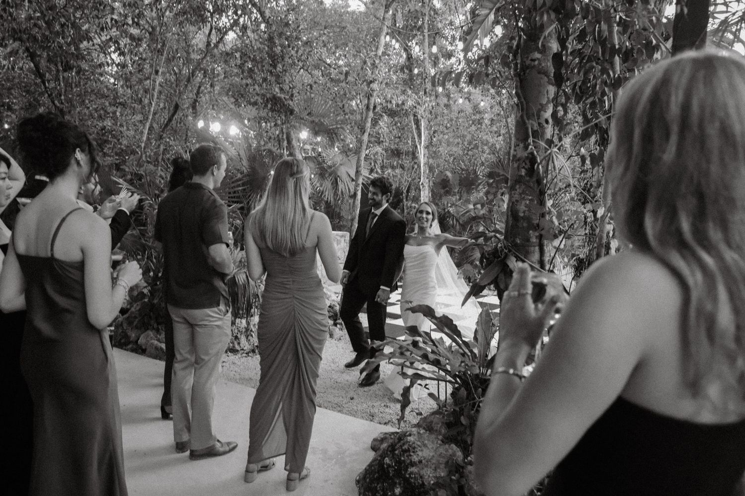 Black and white outdoor wedding ceremony in a forest setting with guests watching the couple exchange vows.