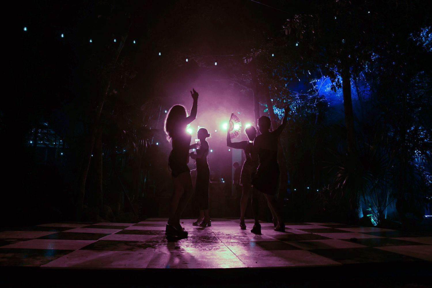 Silhouettes of dancers on a checkered floor bathed in purple and blue stage lighting.