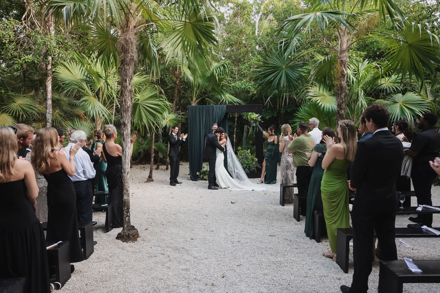 Outdoor wedding ceremony in a tropical garden with guests in black and green attire lining the sandy aisle.