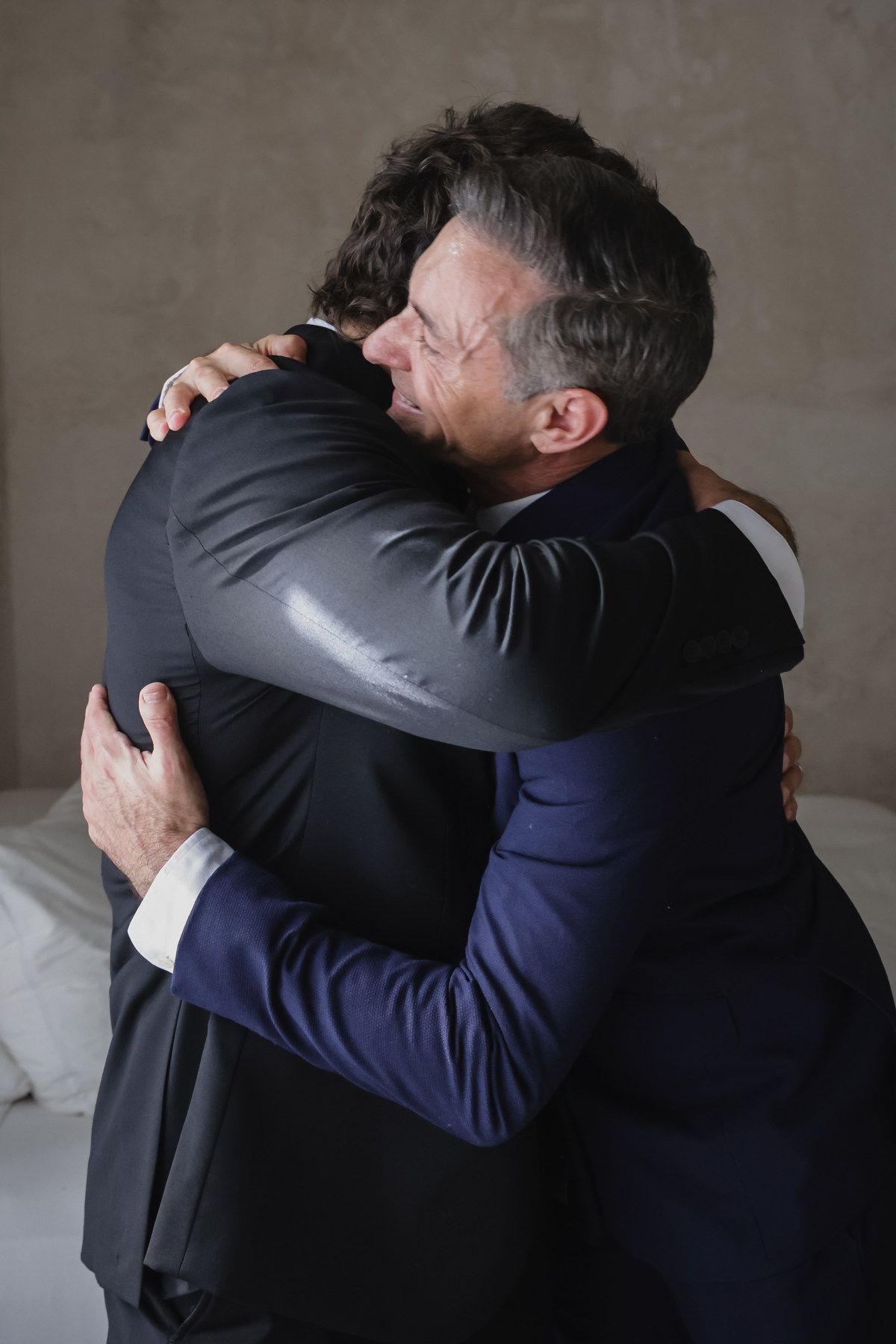 Two men in suits share a warm embrace, one wearing glasses, in an emotional wedding day moment.