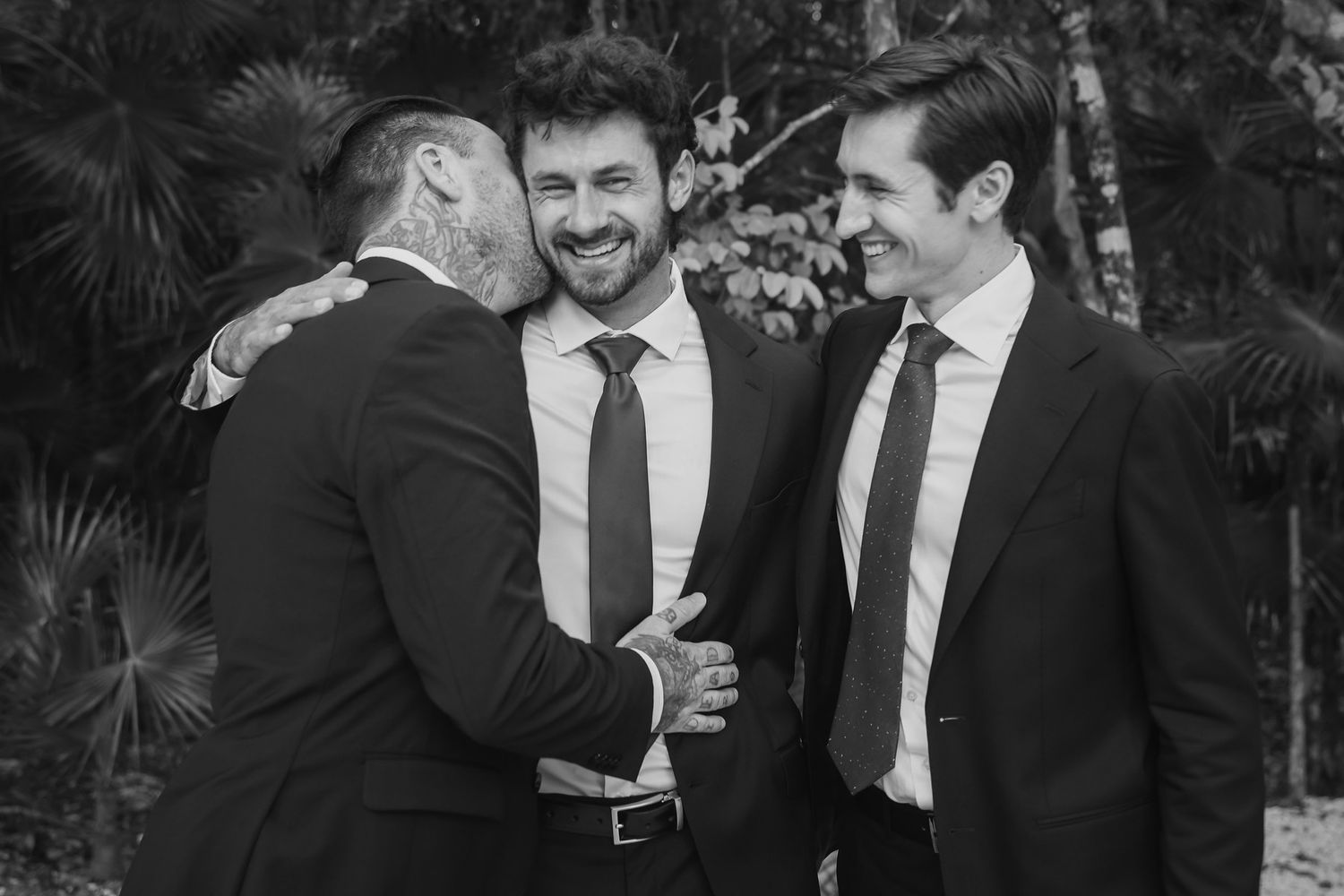 Three men in dark suits share a joyful moment, laughing and embracing outdoors in a black and white photo.