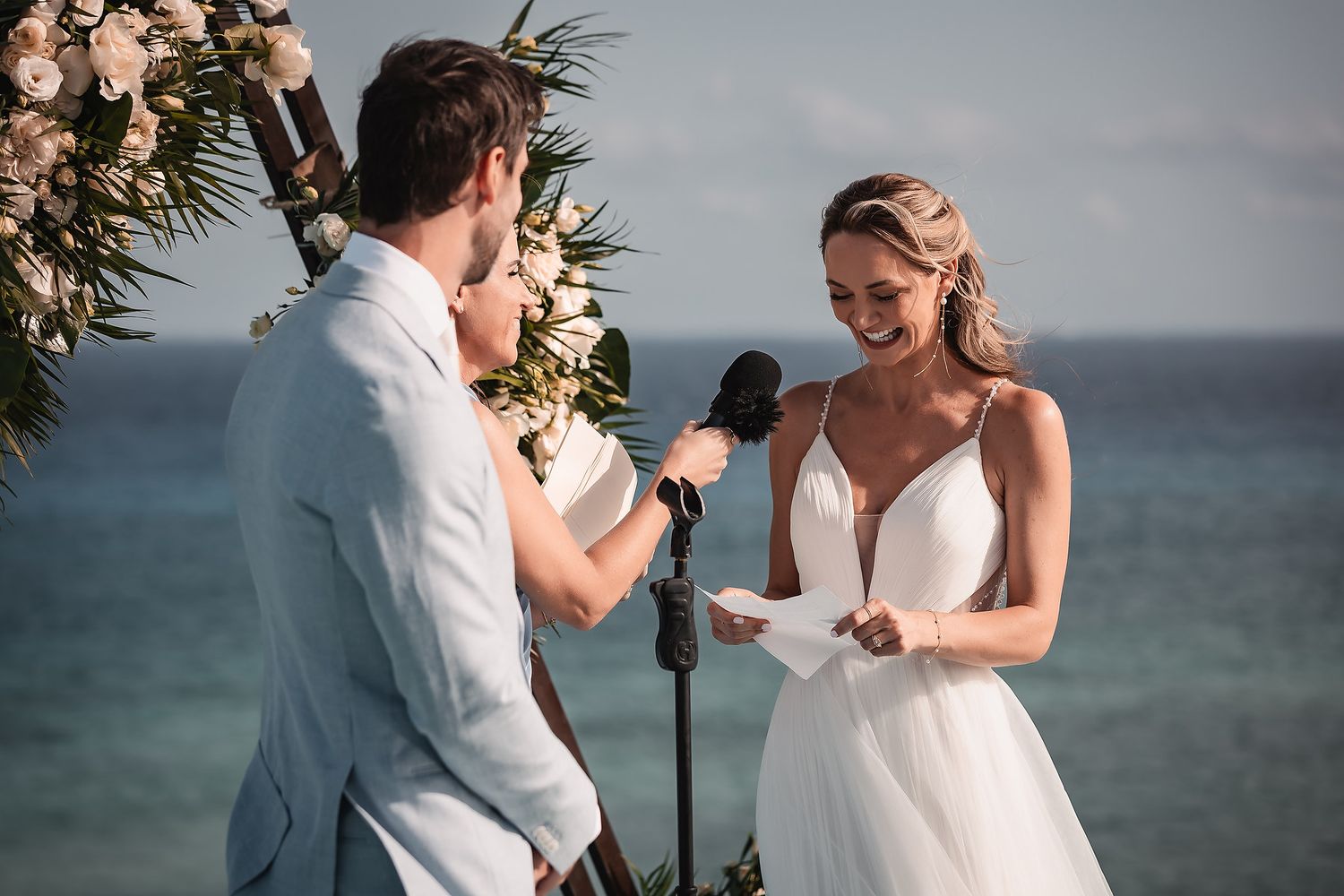 Couple exchanging vows during oceanside ceremony with floral triangle arch and microphone.