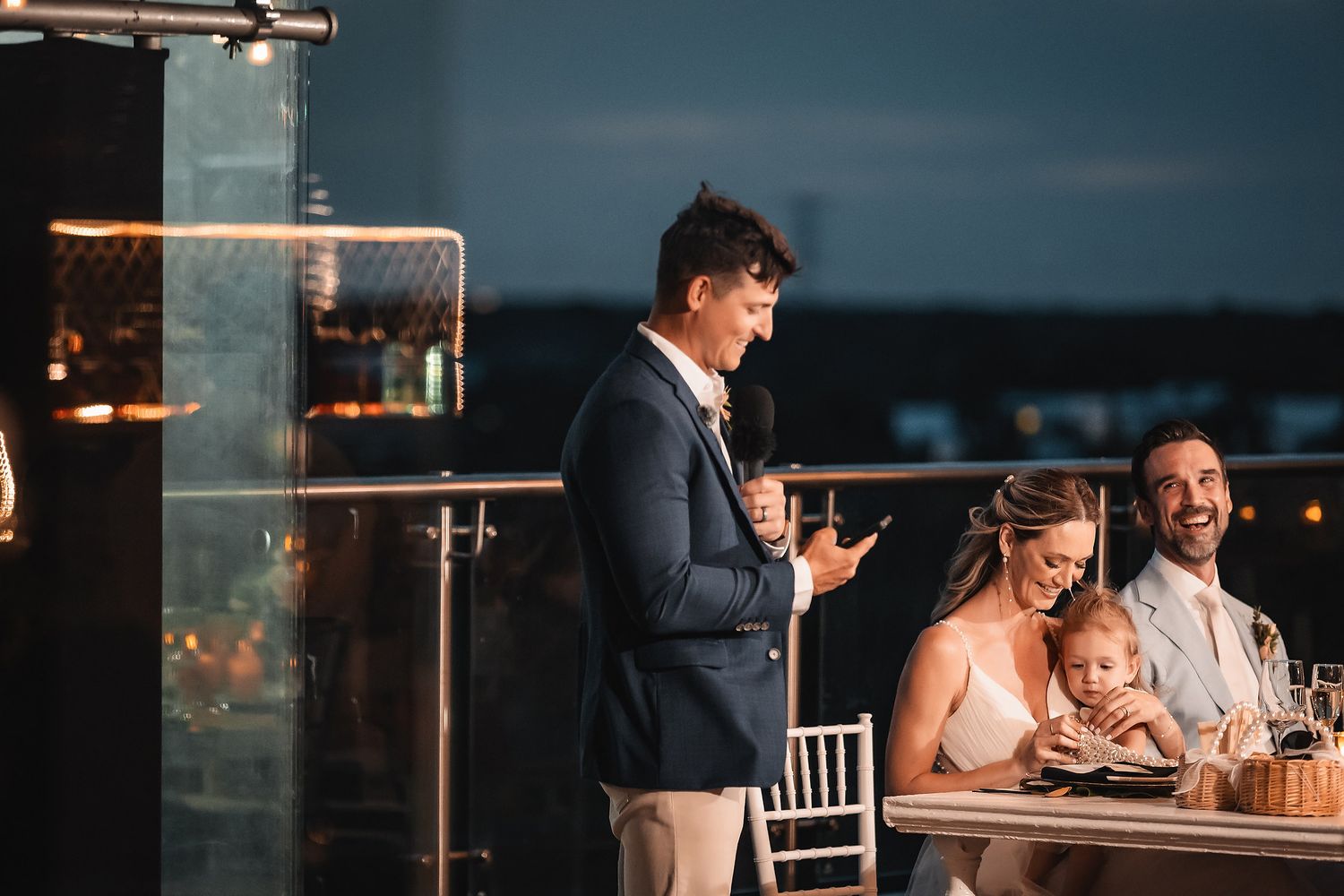 Evening wedding reception with speaker giving toast at oceanfront venue with city lights in background.
