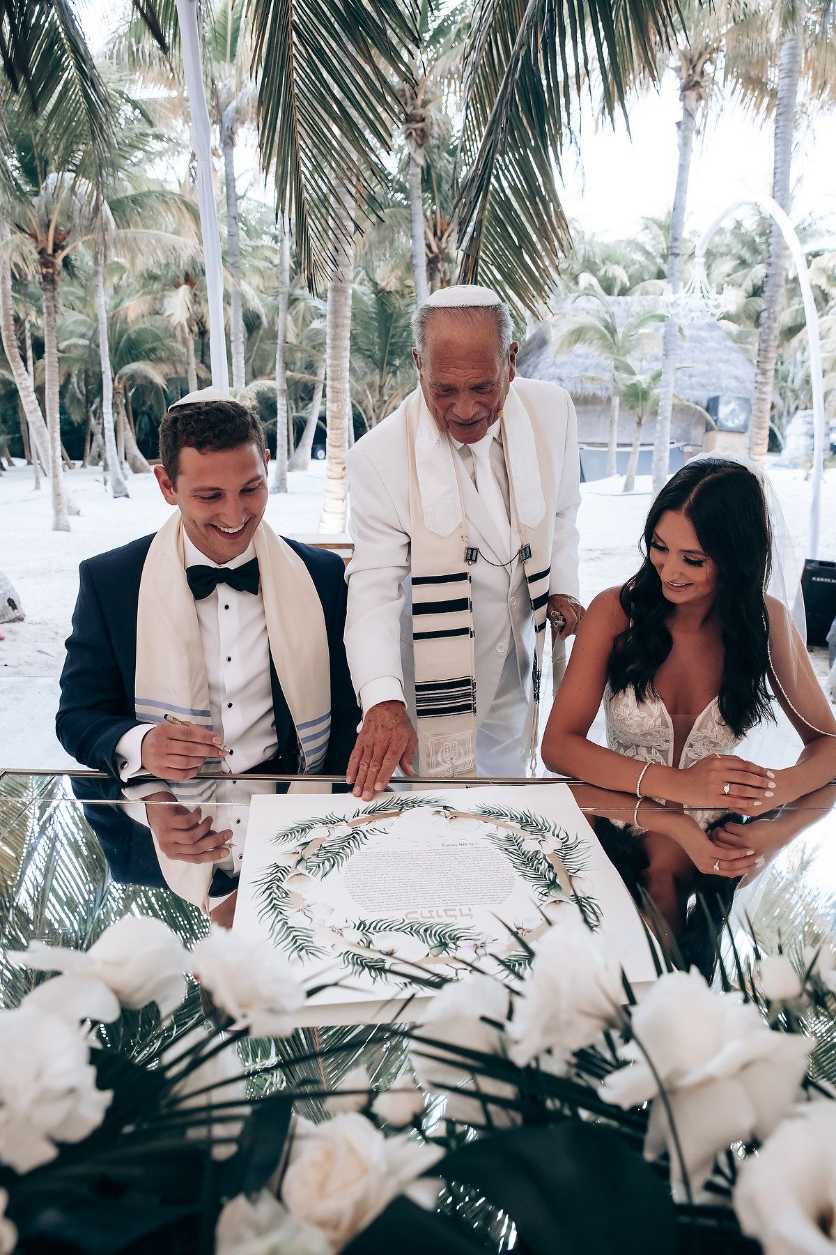 Wedding party members seated at a table with white roses and palm trees in the background during a tropical ceremony.