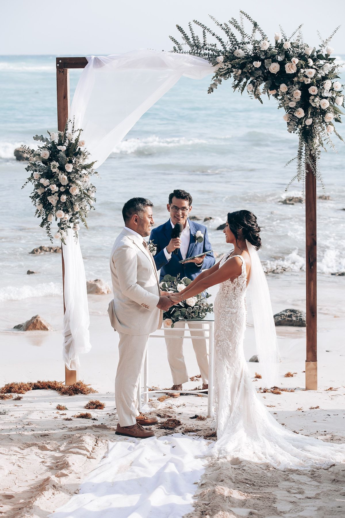 A romantic beach wedding ceremony setup with wooden arch and white floral arrangements along the ocean shore.